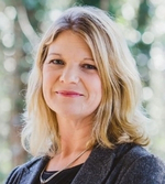 A woman with shoulder-length blonde hair, wearing a dark blazer over a black top, stands outdoors with a blurred background of green trees and sunlight. She is smiling gently at the camera.