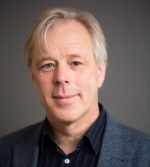 A middle-aged man with light hair and fair skin, wearing a blue blazer over a dark collared shirt, looks at the camera with a slight smile. The softly lit, plain background gives this CUP Global professional a polished and approachable appearance.