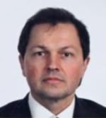 A man with short dark hair, wearing a white shirt, dark jacket, and tie, poses for a formal portrait against a plain light background.