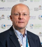 A middle-aged man wearing glasses and a suit jacket with a lanyard stands in front of a backdrop featuring various logos and text.