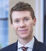 A man with short, light brown hair wearing a dark suit, white shirt, and striped tie, smiling slightly, stands indoors with blurred windows in the background.