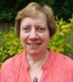 A woman with short light brown hair smiles at the camera. She is wearing a coral-coloured top and standing outdoors in front of green and yellow foliage.