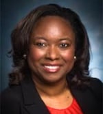 A woman with shoulder-length dark hair smiles, wearing a black blazer over a red top, posed against a dark blue background.