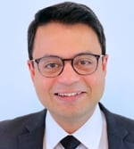 A man wearing glasses, a dark suit, white shirt, and dark tie, smiling in front of a plain light blue background.