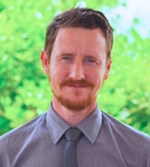 A man with short brown hair, a trimmed beard and moustache, wearing a grey collared shirt and tie, stands outdoors with green leafy trees in the background.