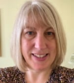 A woman with straight, shoulder-length blonde hair and a fringe smiles at the camera. She is wearing a patterned top and is standing indoors against a light-coloured background.