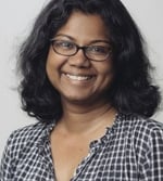 A woman with medium-length curly black hair and glasses smiles at the camera. She is wearing a black and white chequered shirt with a frilled collar. The background is plain and light-coloured.