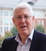 A man with short white hair and glasses is wearing a dark blazer and a chequered shirt, standing outdoors with a blurred building and greenery in the background.