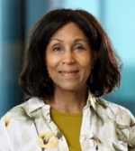 A woman with shoulder-length dark hair smiles softly. She is wearing a floral-patterned shirt over a yellow top, standing in front of a blurred blue and turquoise background at the CUP Global event.