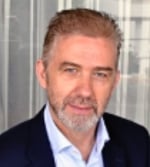 A middle-aged man with light skin, short brown hair, and a neatly trimmed grey beard, wearing a navy blazer and light blue shirt, is sitting in front of a light-coloured curtain.