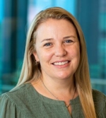 A woman with long blonde hair, wearing a green top, smiles at the camera. The background is softly blurred with shades of blue and teal, suggesting an indoor setting.