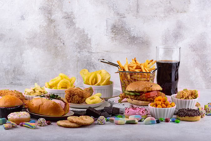 A variety of fast food and snacks, including burgers, chips, fried chicken, crisps, biscuits, colourful sweets, pastries, and a glass of cola, arranged on a light surface against a grey background.