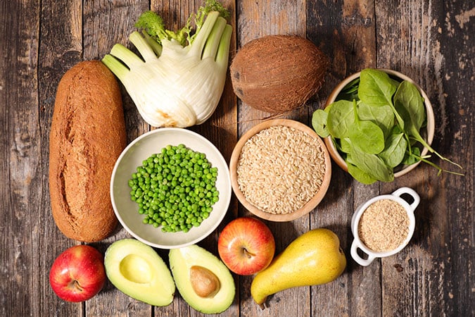 A variety of healthy foods on a rustic wooden table, including wholegrain bread, fennel, coconut, spinach, green peas, brown rice, apples, avocado, pear, and a small bowl of grains.