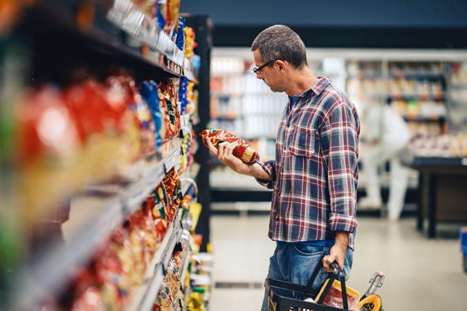 A man wearing glasses and a checked shirt stands in a supermarket aisle, holding a shopping basket in one hand and examining a bag of crisps in the other. Shelves stocked with snacks line both sides of the aisle.