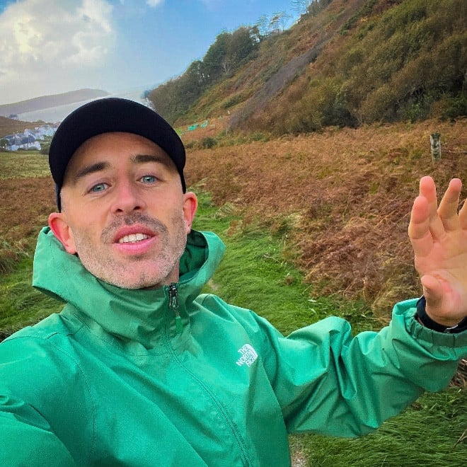 A person in a green jacket and black cap takes a selfie outdoors, standing on a grassy path with hills and autumn-coloured foliage in the background under a partly cloudy sky.