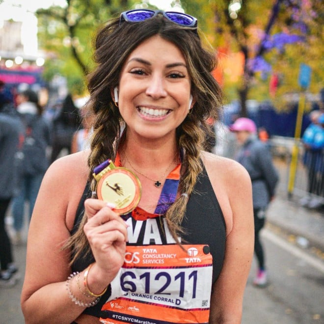 A smiling woman with braided hair holds up a medal after finishing a race. She wears a race number bib numbered 62121 and stands outdoors with other people and trees in the background.