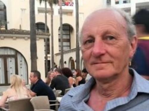 An older man with short grey hair and a blue shirt sits outdoors at a busy café or restaurant, with several people and white buildings in the background.