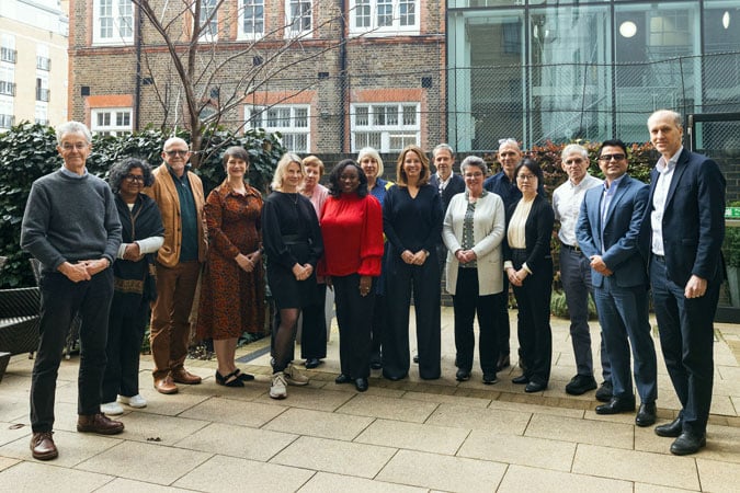 A group of sixteen Global Cancer Update Programme Experts, dressed in business attire, stand outdoors in a courtyard, smiling at the camera. Behind them are brick and glass buildings and leafy greenery.