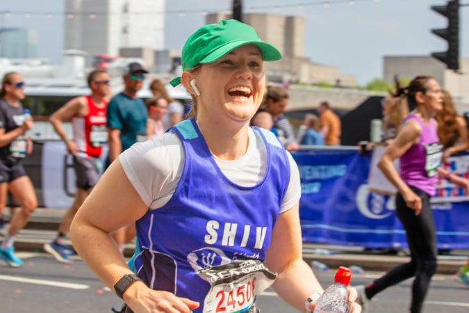 A smiling woman wearing a green cap, blue running vest, and race number runs in a marathon, holding a water bottle—showcasing the energy and spirit that embody the UK Strategy 2026–2030. Other runners and spectators are visible in the background on a sunny day.