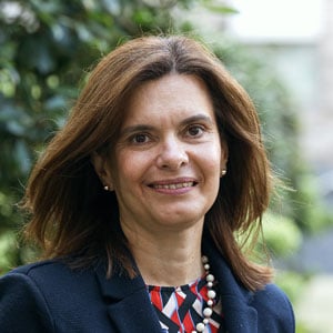 A woman with straight brown hair, wearing a dark blazer, colourful patterned top, pearl necklace, and small earrings, smiles outdoors with greenery in the blurred background.