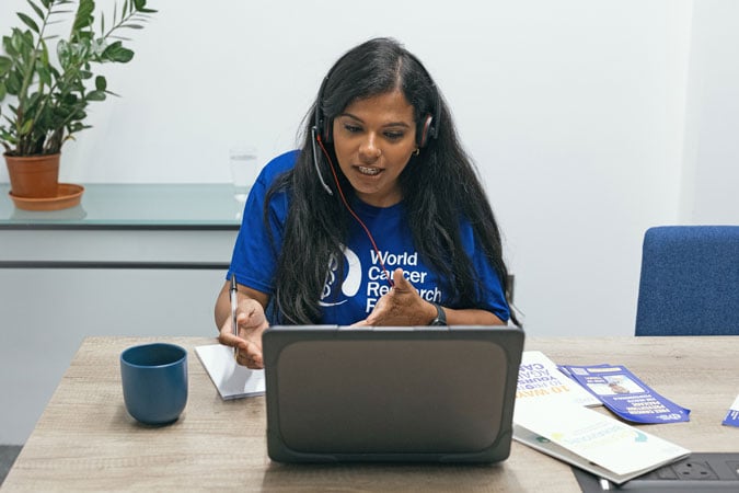 A woman with long dark hair, wearing a blue World Cancer Research Fund T-shirt and a headset, speaks during a video call about cancer prevention workshops for health professionals at a desk with a laptop, notepad, mug, leaflets, and a plant in the background.