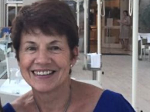 A woman with short brown hair and a blue top smiles at the camera while sitting outdoors at a restaurant. There are tables, chairs, and another person in the background.