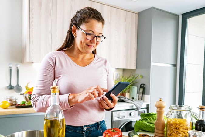 A woman wearing glasses and a light pink shirt uses her mobile in a modern kitchen, surrounded by fresh vegetables, pasta, olive oil, and seasonings on the worktop—planning meals inspired by the UK Strategy 2026–2030.