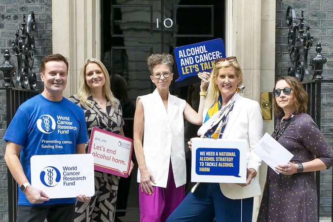 Five people stand outside a building with a door marked 10, holding signs about alcohol and cancer awareness. Their messages, like “Let’s end alcohol harm,” support the UK Strategy 2026–2030 in tackling alcohol-related health risks.