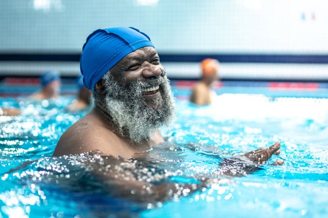 An older man with a grey beard and blue swim cap smiles joyfully whilst swimming in an indoor pool, reflecting the vibrant spirit at the heart of the UK Strategy 2026–2030. Other swimmers are blurred in the background.