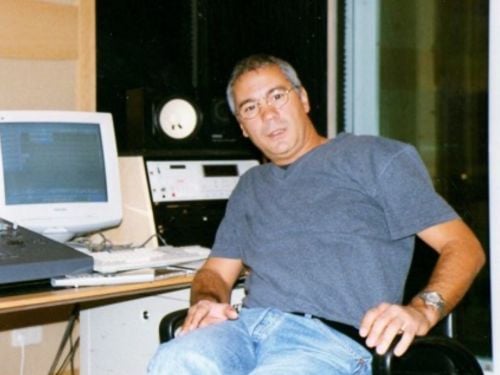 A man wearing glasses and a grey t-shirt sits on a chair in a recording studio with audio equipment, a computer monitor, and speakers visible in the background.