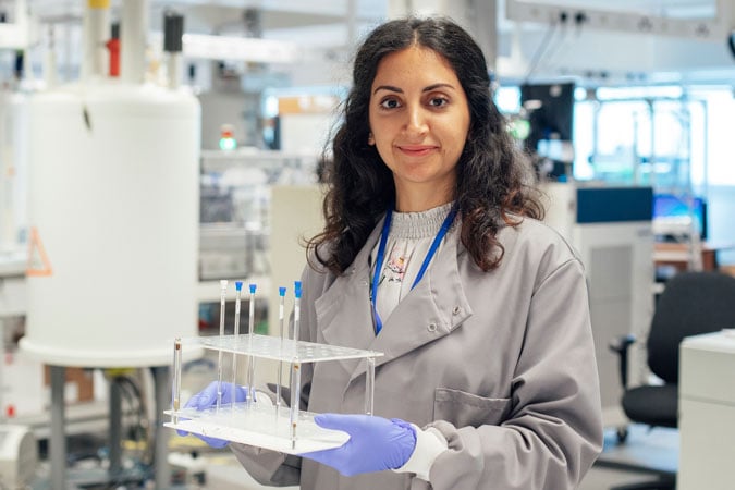 A woman in a lab coat and gloves smiles whilst holding a plastic rack with test tubes in a bright laboratory filled with scientific equipment, reflecting the innovative spirit of the UK Strategy 2026–2030.
