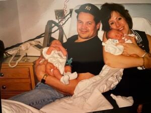 A man and woman sit on a bed, each holding a newborn baby. The man wears a dark baseball cap, the woman is smiling, and there is a phone and lamp on the bedside table behind them.