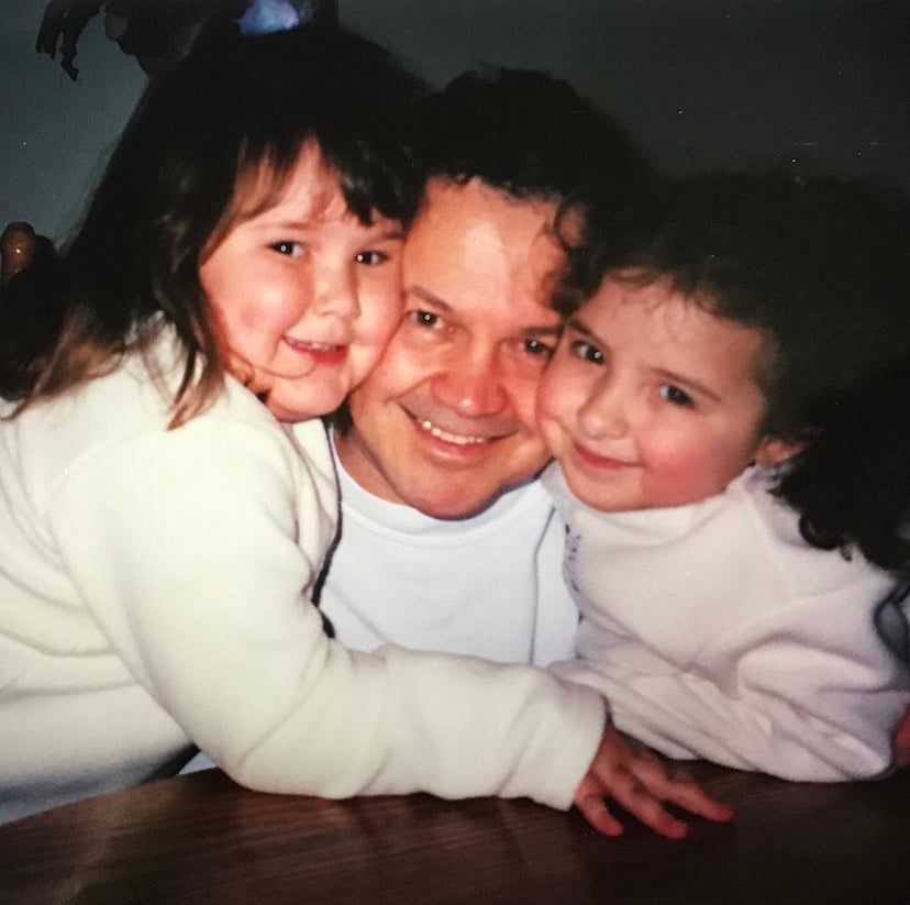 A smiling man sits at a table with two young girls hugging him on each side. All three are wearing light-coloured clothes and look happy and close together.