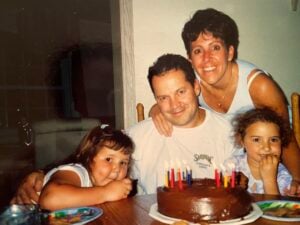 A man sits at a table with a chocolate birthday cake topped with candles, surrounded by two young girls. A woman stands behind him, smiling with her arms round him. Everyone looks happy and festive.
