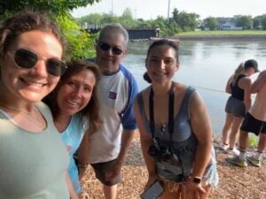 Four people smile for a selfie outdoors by a river, with trees and sunlight in the background. Two other people stand near the water, looking away. The group appears to be enjoying a casual day out.