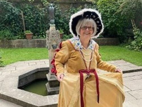 An older woman smiles outdoors, wearing a long yellow historic-style dress, a decorated bonnet, and holding her skirt out. She stands on stone paving near a small fountain with greenery in the background.