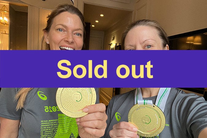 Two women wearing grey shirts and holding medals smile for a selfie indoors. A bold purple banner with yellow text reading Sold out covers the middle of the image.