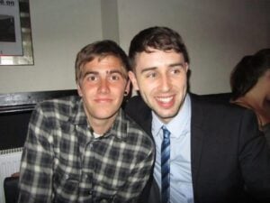 Two young men are sitting closely together indoors. One wears a checked shirt and has an earring, while the other is dressed in a suit and tie, smiling widely. The background has a white wall and part of another person is visible.
