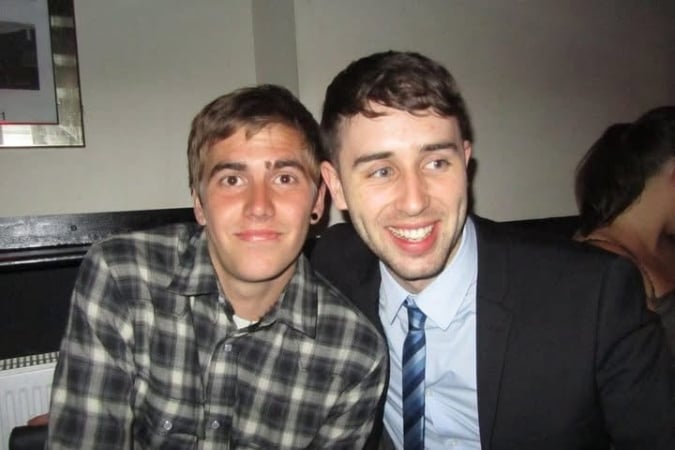Two young men are sitting close together, smiling at the camera. One is wearing a checked shirt with a black earring, and the other is in a suit and tie. They appear to be in an informal indoor setting.