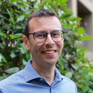  A man with short dark hair and glasses, wearing a light blue collared shirt, smiles whilst standing outdoors in front of green leafy plants. Steven Greenberg, UK Director 