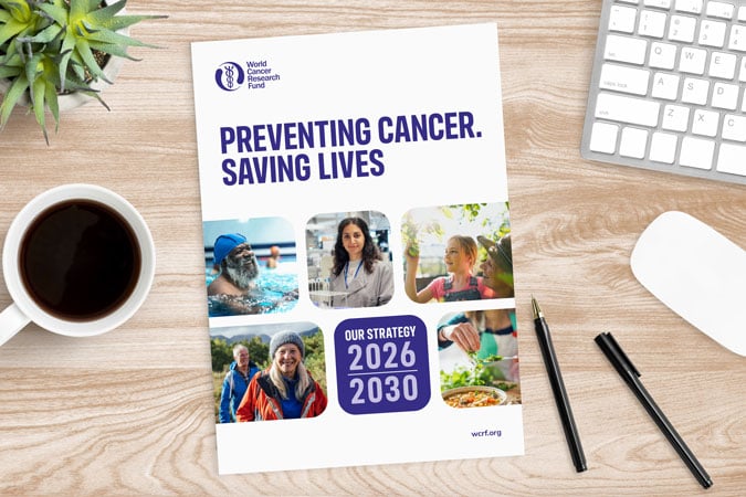 A brochure on a desk titled Preventing Cancer. Saving Lives with photos of diverse people, a coffee cup, plant, keyboard, mouse, and pens beside it. The brochure notes Our Strategy 2026-2030.