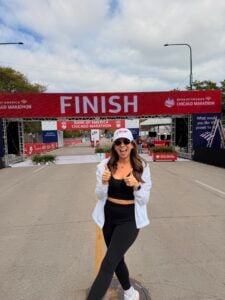A woman in sportswear smiles and gives two thumbs up while standing in front of the Chicago Marathon finish line banner on a cloudy day.