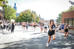 Runners participate in a marathon on a city street lined with trees and spectators. A smiling woman in the foreground runs towards the camera, wearing sunglasses and holding up a peace sign. Other runners and tall buildings are visible in the background.