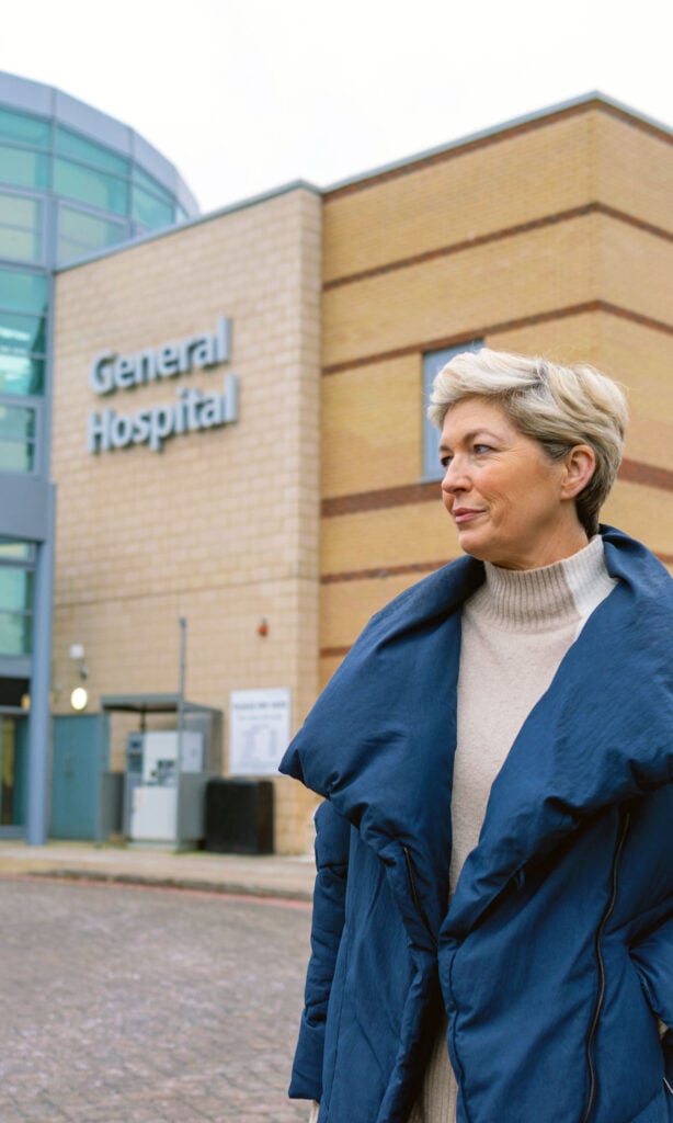 A woman with short grey hair and a blue coat stands outside a building with a sign that reads General Hospital.
