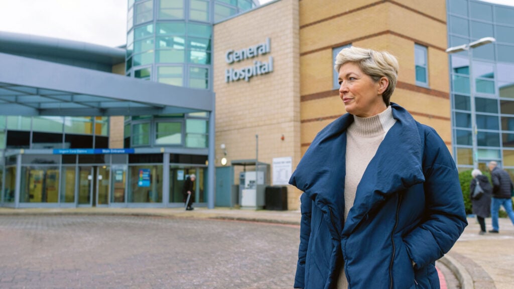 A woman with short grey hair, wearing a blue coat, stands outside the entrance of a modern hospital building labelled General Hospital. Other people are visible near the entrance in the background.