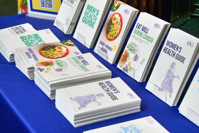 A table displays stacks of health guides, including “Men’s Health Guide,” “Women’s Health Guide,” and “Eat Well During Cancer,” all arranged neatly on a royal blue tablecloth, promoting awareness about movement and sedentary behaviour cancer risk.