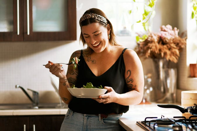 A woman with tattoos smiles whilst eating a salad in a bright kitchen. She wears a black vest, jeans, and a headband, standing near the cooker and sink with plants and dried flowers in the background.