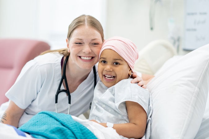 A smiling nurse with a stethoscope hugs a young girl wearing a headscarf, sitting up in a hospital bed. Both are smiling and looking at the camera in a bright hospital ward.
