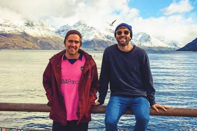 Two men smiling in front of a scenic lake with snow-capped mountains in the background. One wears a beanie and pink shirt, the other sunglasses and a dark jumper. A bird flies above them in the partly cloudy sky.