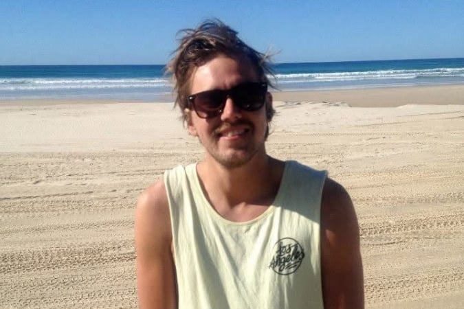 A man wearing sunglasses and a sleeveless light shirt stands on a sandy beach with the sea and clear blue sky in the background.